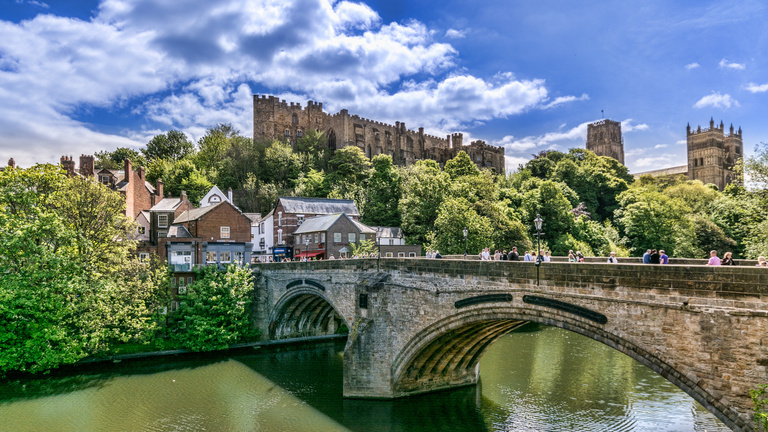 Stone bridge over a calm river, overlooked by a historic castle and cathedral, set amid lush green trees under a vibrant blue sky with scattered clouds.
