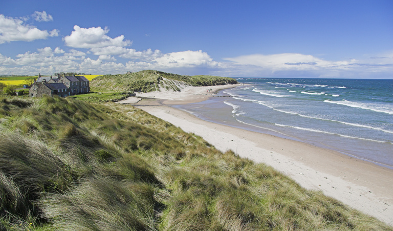 A serene coastal landscape featuring a sandy beach with gentle waves, grassy dunes in the foreground, and a rustic stone house on the left under a blue sky.