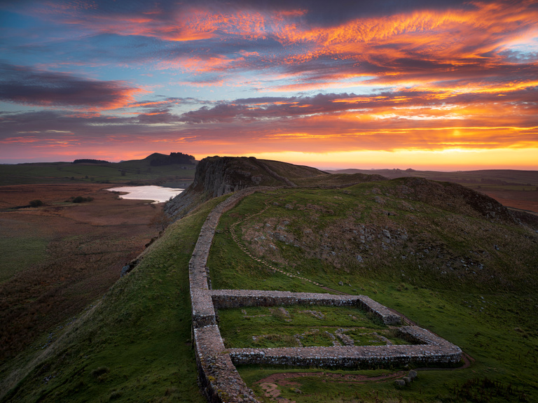 Sunset over Hadrian's Wall with vibrant orange and purple sky. The ancient stone wall winds over green hills, evoking a sense of history and tranquility.