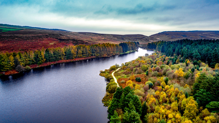 A vibrant aerial view of a serene river flanked by colorful autumn trees. A winding path follows the riverbank, under a blue and cloudy sky.
