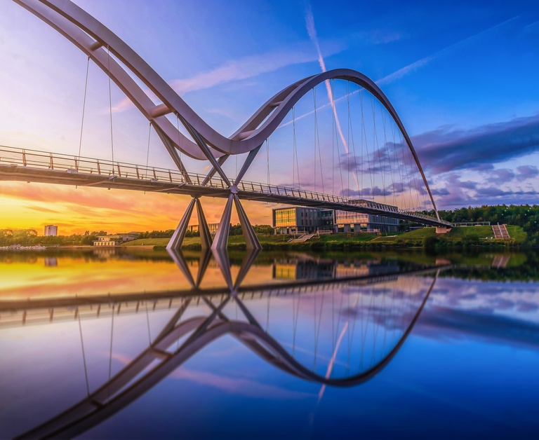 A modern, curved pedestrian bridge spans a calm river at sunset, reflecting on the water. The sky is vibrant with blue, purple, and orange hues.